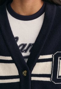 Granatowy kardigan w białe paski, z dekoltem w serek i pojedynczym zapięciem na guzik. Nosi się go na jasnoszarym T-shircie z niebieskimi detalami.