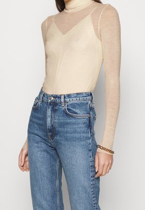 Beige, durchsichtige Langarmbluse mit Stehkragen, kombiniert mit blauen, hoch geschnittenen Jeans. Auffälliges goldenes Kettenarmband am Handgelenk.