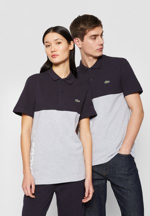 Dos personas llevan camisetas polo de manga corta con una parte superior azul marino y una inferior gris, con un pequeño logo verde en el pecho.