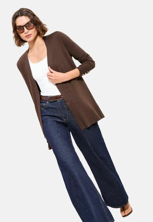 Woman wearing brown open cardigan, white tank top, wide-leg blue jeans, brown belt, sunglasses, and brown sandals, posing against white background.