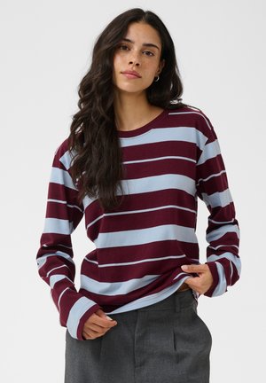 Jeune femme aux longs cheveux foncés portant un t-shirt à manches longues rayé bordeaux et bleu clair ainsi qu'un pantalon gris, debout devant un fond uni.