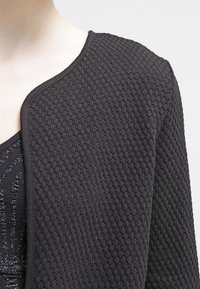 Czarna, strukturalna kurtka z wzorem diamentowym, zaokrąglonym dekoltem i gładkimi krawędziami. Zbliżenie na detale i strukturę tkaniny.