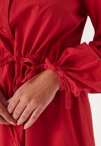 Abito rosso con bottoni, vita elastica, polsini a maniche gather con nastri in stoffa, texture di cotone morbido.