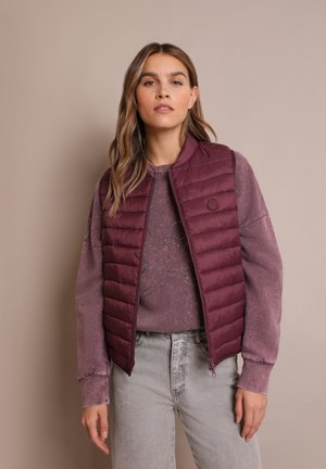 Bordeaux puffer gilet met horizontale quiltpatroon, ronde hals en een klein logo detail, gelaagd over een paarse sweater en grijze spijkerbroek.