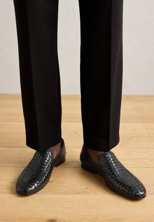 Zwarte gevlochten leren instappers met een gladde, glanzende afwerking en een laag profiel, gecombineerd met zwarte maatwerkbroek op een houten vloer.