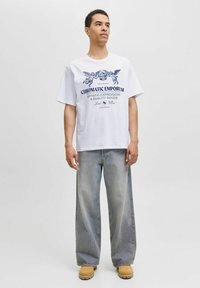 Weißes Kurzarm-T-Shirt mit blauem Grafikdruck, das Engelchen und Text zeigt, kombiniert mit hellblauen weit geschnittenen Jeans und beigen Schuhen.