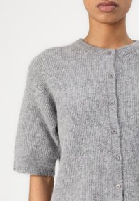 Gros plan d'une personne portant un cardigan en maille douce, gris, à manches courtes, boutonné, avec un col rond sur un fond uni.