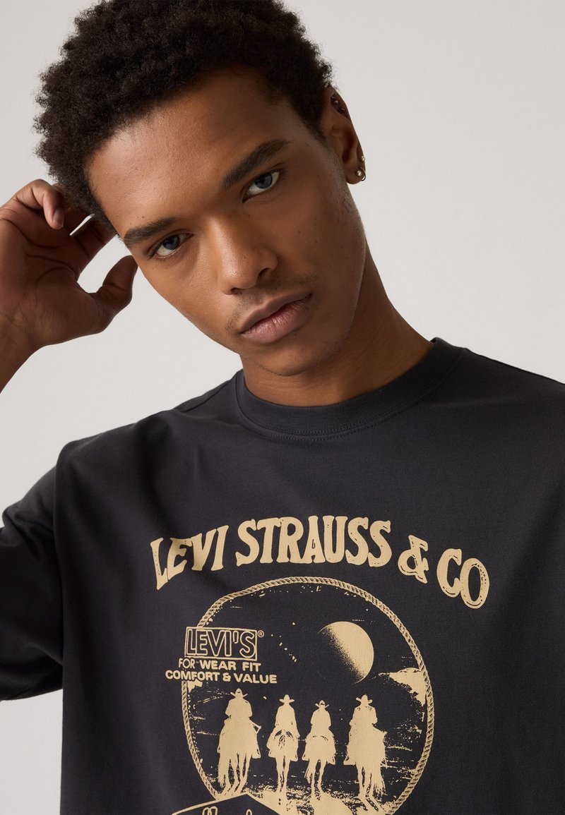 Schwarzes T-Shirt mit kurzen Ärmeln, das ein Retro-Design mit Cowboys und in cremefarbener Schrift zeigt. Aus weichem Baumwollmaterial und mit Rundhalsausschnitt.