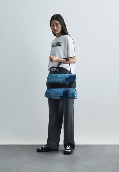 Νεαρή γυναίκα με λευκή μπλούζα Moschino και μαύρο παντελόνι, κρατώντας μια μεγάλη μπλε τσάντα από υφασμάτινη καπιτονέ με μαύρη λαβή, στέκεται σε γκρι δάπεδο.