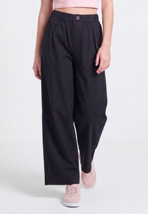 Person trägt lockere schwarze Hose mit elastischem Bund und Knopf, kombiniert mit hellrosa Sneakern und einem kurzen pinken Oberteil.