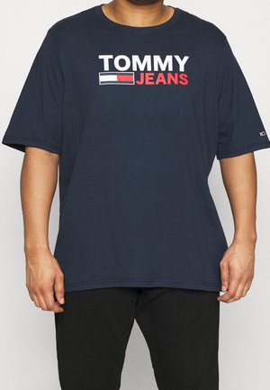 Uomo che indossa una T-shirt Tommy Jeans blu navy con logo bianco e rosso, abbinata a pantaloni neri su sfondo semplice.