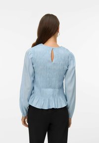 Šviesiai mėlyna palaidinė su raukšlėtu nugarėle, ilgomis rankovėmis ir raktelio formos iškirpte prie kaklo. Turi išplatėjusį apatinį kraštą ir glotnią, švelnią tekstūrą.