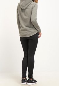 Persona che indossa un felpa grigia con cappuccio, jeans skinny neri e scarpe da ginnastica nere Nike, in piedi con la schiena rivolta verso l'osservatore su uno sfondo neutro.