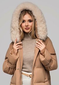 Giacca puffer beige con un ampio cappuccio in pelliccia sintetica, caratterizzata da una texture trapuntata e una zip frontale. Indossata sopra un maglione beige a collo alto.