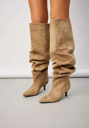 Bottes beiges en daim souples montant jusqu'au genou, avec des bouts pointus et de petits talons kitten, portées sur des jambes nues.