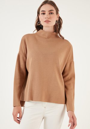 Maglione lavorato a maglia di un marrone chiaro, con collo alto e maniche scese. L'orlo è leggermente arrotondato, abbinato a jeans bianchi.