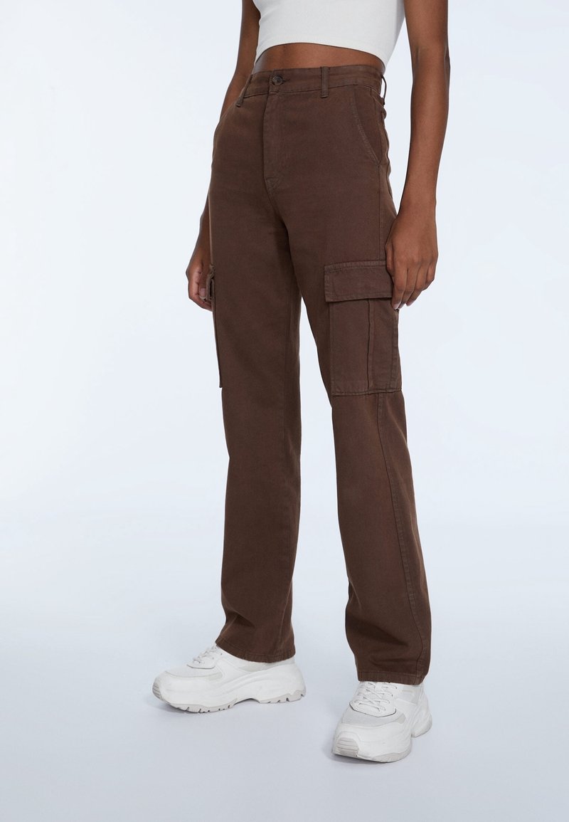 Bruine cargobroek met zijzakken, gemaakt van stevig materiaal. Heeft een recht model en een hoge taille. Gedragen met witte sneakers.