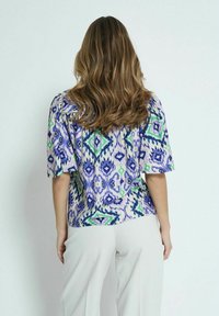 Blusa ampia realizzata in tessuto stampato con forme a diamante di colore viola, verde e blu. Maniche scese e orlo arrotondato.