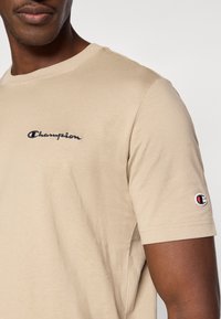 Μπεζ βαμβακερό t-shirt με στρογγυλή λαιμόκοψη, με κεντημένο λογότυπο «Champion» σε ναυτικό μπλε στο στήθος και διακοσμητικό λογότυπο  στο μανίκι.