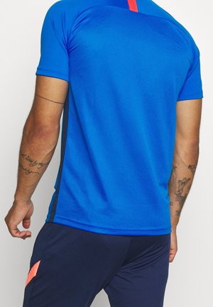 Homme portant un maillot de sport bleu vif à manches courtes avec un short bleu foncé et corail, montrant des bras tatoués, debout sur un fond blanc.