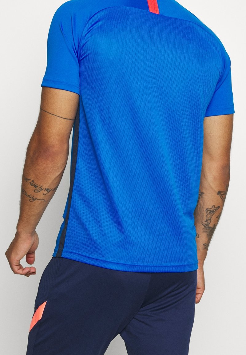 Homme portant un maillot de sport bleu vif à manches courtes avec un short bleu foncé et corail, montrant des bras tatoués, debout sur un fond blanc.
