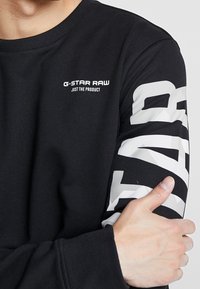 Černá mikina s žebrovaným výstřihem, bílým logem G-Star RAW a přeplněným bílým grafikou na rukávu. Měkký materiál, dlouhé rukávy.