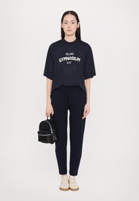 Marinblå mesh t-shirt med tryck "MILANO GYMNASIUM", matchad med marinblå tapered byxor och en liten svart ryggsäck. Beiga skor med ljusa snören.