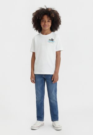 Barn med krøllet hår iført hvid Converse T-shirt, blå jeans og hvide sneakers, stående mod en ensfarvet baggrund.