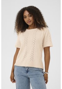 Ženska s kodrastimi lasmi nosi svetlo breskvasto kratkoprstno pleteno majico s chevron vzorcem in modre kavbojke, stoji ob nevtralni ozadju.