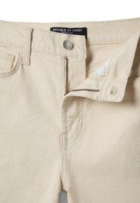 Jeans beige de corte bota con cremallera frontal parcialmente abierta, cierre con botón metálico y etiqueta "Republic of Denim" en la cintura.