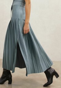 Plissé lichtblauwe maxi-rok met een zijsplit en bijpassende riem, gecombineerd met zwarte laarzen met hak, die een egaal textuur en vloeiende beweging tonen.