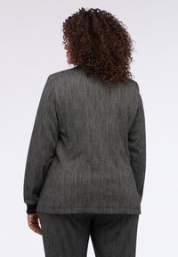 Ušitý šedý saka s texturovaným povrchem, s jednoduše zapínáním a černými manžetami, zobrazeno zezadu.