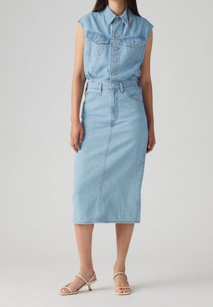 Frau trägt ärmelloses, hellblaues Jeans-Top mit Knöpfen, in einen passenden hoch taillierten Midi-Jeansrock gesteckt, dazu beige Riemchensandalen.