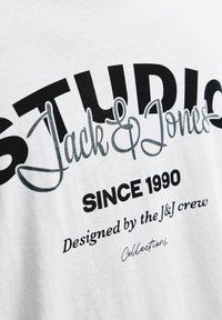 Hvid bomulds T-shirt med sort og teal grafisk tekst: "STUDIO Jack & Jones" og "Siden 1990" i forskellige skrifttyper. Glat tekstur.