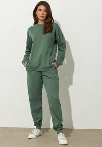 Sudadera de algodón verde y joggers a juego con un corte relajado, que cuentan con puños acanalados y bolsillos, combinados con zapatillas blancas.