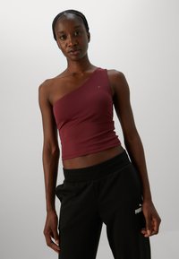 Vrouw in een bordeaux rozen één-schouder crop top en zwarte Puma joggingbroek, staande tegen een effen lichte achtergrond.