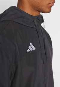 Giacca nera con cappuccio e pannello frontale testurizzato, con logo Adidas grigio. Dotata di coulisse regolabile e finitura in tessuto liscio.