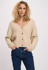Beżowy sweter z dzianiny z głębokim dekoltem w literę V, sześcioma brązowymi guzikami, prążkowanymi mankietami i w pasie. Nosi się go na ciemnoniebieskie dżinsy.