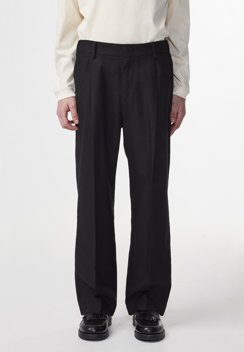 Man met zwarte op maat gemaakte broek en zwarte leren instappers met franjes, in combinatie met een off-white langwerpige top.