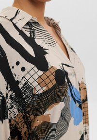 Chemise collerette avec un motif abstract composé de formes irrégulières noires, bleues et brunes sur un tissu beige, mettant en valeur des détails de boutons.