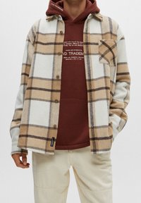 Mand iført en beigefarvet og brunternet knap-shirt over en rustfarvet hoodie med tekst, parret med off-white bukser, står med hænderne i lommerne.