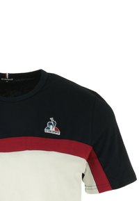 T-shirt a maniche corte in tessuto blu navy, con una striscia orizzontale rossa e un pannello bianco con logo sul lato sinistro.
