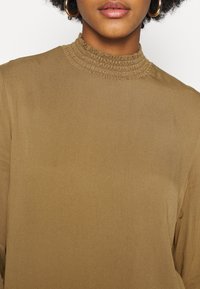 Maglione marrone con collo alto e tessuto arricciato nella zona del collo, realizzato in materiale morbido. Presenta maniche lunghe e una vestibilità rilassata.