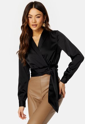 Blusa avvolgente in satin nero con maniche lunghe, scollatura a V profonda e dettaglio sulla vita annodato. Abbinata a pantaloni beige in ecopelle a vita alta.