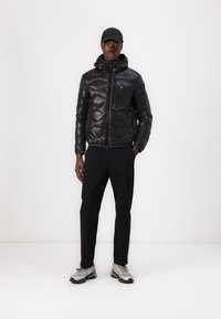 Giacca a vento nera trapuntata con cappuccio e chiusura con zip, dotata di tasca sul petto e sottili dettagli verdi, abbinata a pantaloni neri e scarpe grigie.