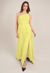 Einseitiges Abendkleid aus leuchtend gelbem Stoff, mit Falten, einem seitlichen Schlitz und einem fließenden Design, ergänzt durch schwarze High Heels.