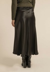 Jupe A-line en satin noir avec une texture lisse, un design fluide, un ourlet à mi-genou, portée avec des bottes noires à hauteur de genou.