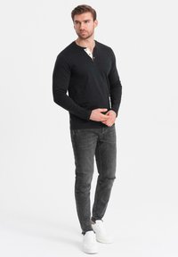 Schwarzes Langarm-Henley-Shirt mit einer Dreiknopf-Leiste und sandfarbenen Akzenten, kombiniert mit grauen, abgewetzten Slim-Fit-Jeans und weißen Sneakers.