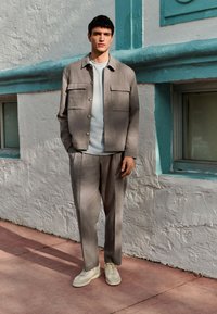 Chaqueta de lino gris claro con dos bolsillos delanteros sobre una camiseta gris, combinada con pantalones a juego y zapatillas crema frente a una pared blanca texturizada.