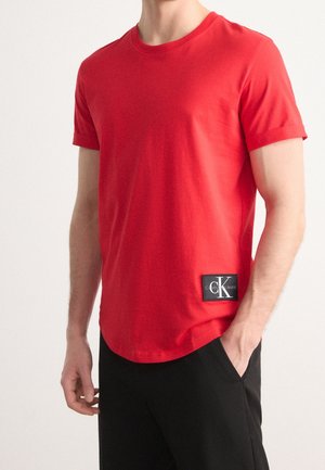 T-shirt en coton rouge avec un col rond, des manches courtes et un patch logo noir en bas à gauche. Texture lisse et coupe ajustée.